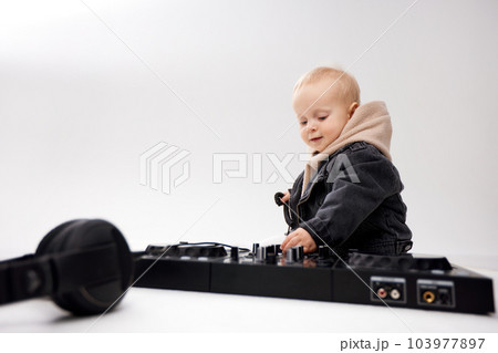 A cute kid in a beige hoodie and a funny black motorcycle jacket sits on the floor with dj headphones and a dj mixing console. Music and fun. Isolate on a white background. 103977897
