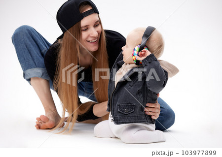 A young barefoot mom in a black top, blue loose jeans and a black baseball cap and her toddler in a black motorcycle jacket are sitting on the floor. The kid has multi-colored headphones on his head. A young barefoot mom in a black top, blue loose jeans and a black baseball cap and her toddler in a black motorcycle jacket are sitting on the floor. The kid has multi-colored headphones on his head. 103977899