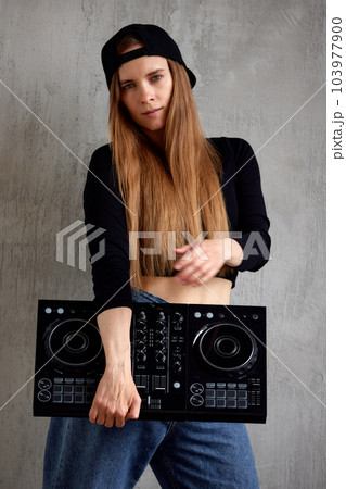 A young pretty long-haired DJ girl in a black top, blue loose jeans and a black baseball cap stands against a gray wall and holds a black DJ mixing console in her hands. Music and fun. A young pretty long-haired DJ girl in a black top, blue loose jeans and a black baseball cap stands against a gray wall and holds a black DJ mixing console in her hands. Music and fun. 103977900