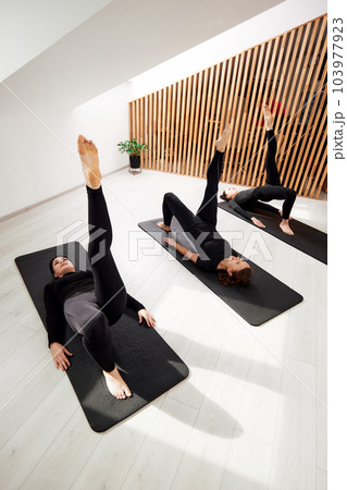 Young women are doing Pilates in a bright studio. Three slender sportswomen in black suits do exercises lying on yoga mats. Healthy lifestyle concept. 103977923