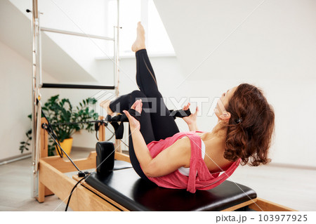 A woman is doing Pilates on a reformer bed in a bright studio. A