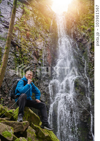 Tourist attraction of Germany - falls of Burgbach Waterfall near Schapbach, Black Forest, Baden-Wurttemberg, Germany. Man hiker in blue jacket standing on stone and looks at flow of falling water 103981567