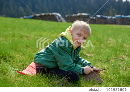 Fun teenager sits on hillside. Boy resting in mountains. School boy sitting of hill in grass field and enjoying beautiful landscape view. Rear view of teenage boy hiker resting in nature. Active 103981601