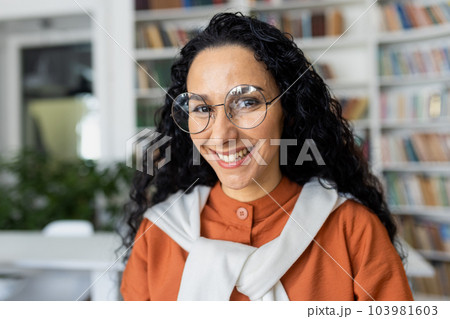 Portrait of young beautiful female student inside university academic library, Hispanic woman with curly hair and glasses close up smiling and looking at camera. 103981603