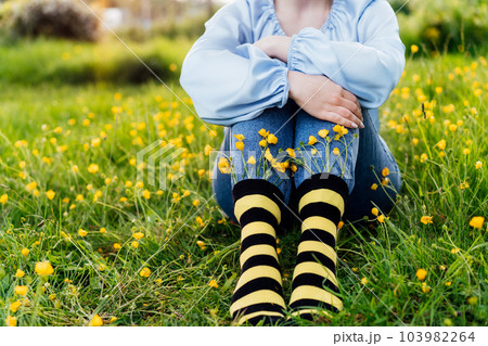 Close up young female wearing jeans and striped black and yellow socks with flowers inside sitting on the green grass of blooming meadow. Concept of bee protection, bloom season, art, creativity 103982264