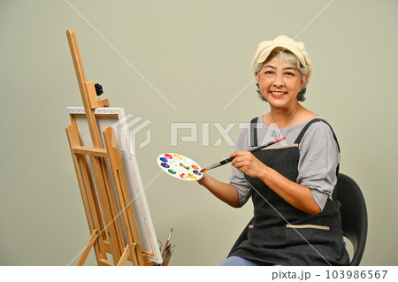 Portrait of beautiful 60s senior woman wearing apron holding brush and palette over gray background Portrait of beautiful 60s senior woman wearing apron holding brush and palette over gray background 103986567