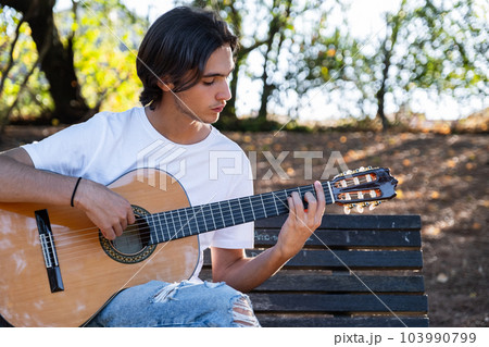 Guitarist plays on guitar outdoors.  Handsome guy plays the guitar on the nature sitting on a bench.  Young man plays a musical instrument outside  house. Musician plays a classical guitar in the 103990799