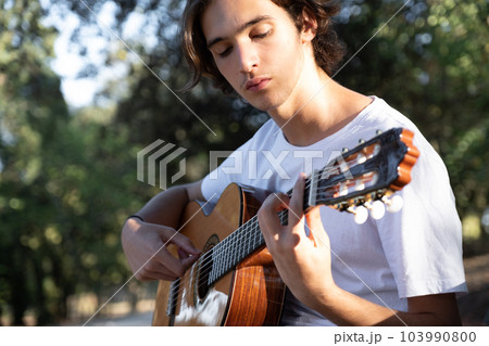 Guitarist plays on guitar outdoors. Handsome guy plays the guitar on the nature sitting on a bench. Young man plays a musical instrument outside house. Musician plays a classical guitar in the Guitarist plays on guitar outdoors. Handsome guy plays the guitar on the nature sitting on a bench. Young man plays a musical instrument outside house. Musician plays a classical guitar in the 103990800