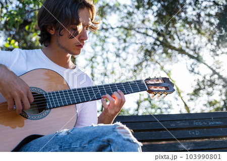 Guitarist plays on guitar outdoors.  Handsome guy plays the guitar on the nature sitting on a bench.  Young man plays a musical instrument outside  house. Musician plays a classical guitar in the 103990801