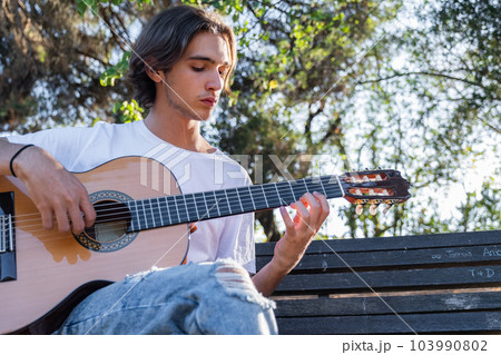Guitarist plays on guitar outdoors. Handsome guy plays the guitar on the nature sitting on a bench. Young man plays a musical instrument outside house. Musician plays a classical guitar in the Guitarist plays on guitar outdoors. Handsome guy plays the guitar on the nature sitting on a bench. Young man plays a musical instrument outside house. Musician plays a classical guitar in the 103990802