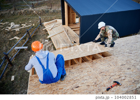 Carpenters mounting wooden OSB board on rooftop of future cottage. Men workers building wooden frame house. Carpentry and construction concept. Carpenters mounting wooden OSB board on rooftop of future cottage. Men workers building wooden frame house. Carpentry and construction concept. 104002851