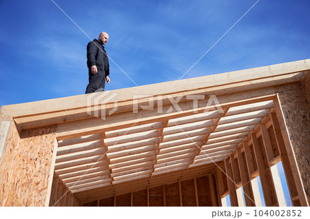 Male designer building wooden frame house. Bald man standing on construction site, inspecting quality of work on sunny day with blue sky on background. 104002852