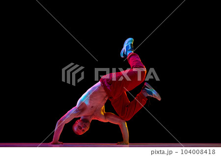 Dance show. Muscular, athletic, shirtless young man dancing breakdance, hip hop against black studio background in neon light. Dance show. Muscular, athletic, shirtless young man dancing breakdance, hip hop against black studio background in neon light. 104008418