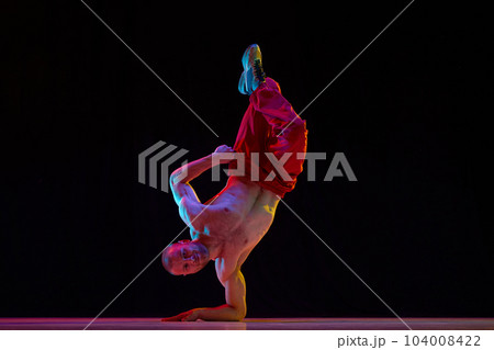 Muscular, strong, shirtless young man dancing breakdance, hip hop against black studio background in neon light 104008422