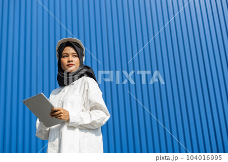 asian muslim woman industrial engineer in uniform wearing safety hard hat using tablet. 104016995