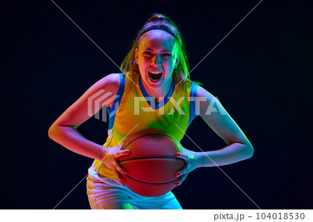Concentrated and motivated young girl, basketball player in uniform posing with ball against black studio background in neon light 104018530