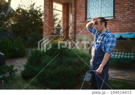 Professional gardener worker in blue working uniform wiping sweat from forehead, cutting green grass with lawn mower, tired after hard work in garden on hot summer day. People Work landscape Gardening 104019415