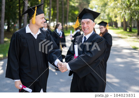 A group of graduates in robes outdoors. An elderly man and a young guy congratulate each other on receiving a diploma. 104023831
