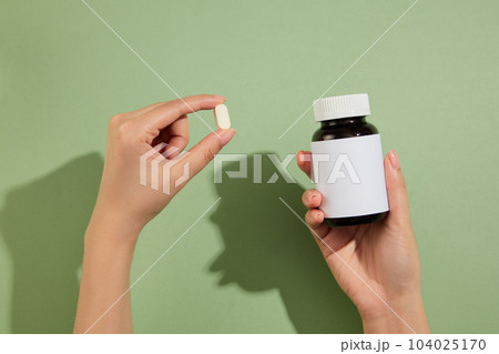 Two hand carrying a small white pill and a blank label medicine bottle. Pastel green background. Mockup of medicine brands 104025170