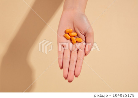 Minimal scene of a hand featuring some orange pills on a beige background. Concept of medicine and health support Minimal scene of a hand featuring some orange pills on a beige background. Concept of medicine and health support 104025179