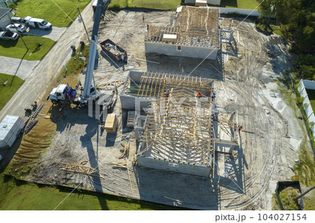 Professional builders and crane truck working on roof construction of unfinished suburban home with wooden frame structure in Florida rural area. Concept of housing development in America Professional builders and crane truck working on roof construction of unfinished suburban home with wooden frame structure in Florida rural area. Concept of housing development in America 104027154
