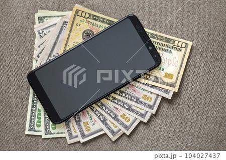 New black modern cellphone on money dollars banknotes background. Modern technology, communication and online trade using gadget concept. New black modern cellphone on money dollars banknotes background. Modern technology, communication and online trade using gadget concept. 104027437