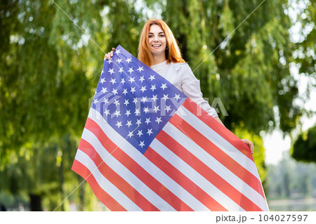 Happy young woman posing with USA national flag standing outdoors in summer park. Positive girl with United States banner outdoors. International day of democracy concept. Happy young woman posing with USA national flag standing outdoors in summer park. Positive girl with United States banner outdoors. International day of democracy concept. 104027597