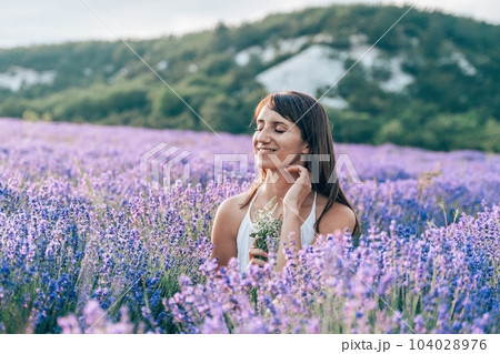 Close up portrait of happy young brunette woman in white dress on blooming fragrant lavender fields with endless rows. Warm sunset light. Bushes of lavender purple aromatic flowers on lavender fields. Close up portrait of happy young brunette woman in white dress on blooming fragrant lavender fields with endless rows. Warm sunset light. Bushes of lavender purple aromatic flowers on lavender fields. 104028976