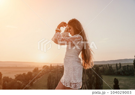 Happy woman in white boho dress on sunset in mountains. Romantic woman with long hair standing with her back on the sunset in nature in summer with open hands. Silhouette. Nature. Sunset. Happy woman in white boho dress on sunset in mountains. Romantic woman with long hair standing with her back on the sunset in nature in summer with open hands. Silhouette. Nature. Sunset. 104029003