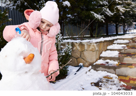 Caucasian lovely little kid girl 5 years old, dressed in pink down jacket and stylish fluffy earmuffs holding a shovel and building a snowman outdoors. Leisure activities and games at wintertime Caucasian lovely little kid girl 5 years old, dressed in pink down jacket and stylish fluffy earmuffs holding a shovel and building a snowman outdoors. Leisure activities and games at wintertime 104043033