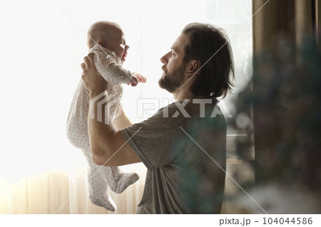 Young neo father is playing with his newborn baby in a nursery morning. Father's Day. Daddy holds in hands little Child and Plays with him. Concept of children, baby, parenthood, childhood, family. Young neo father is playing with his newborn baby in a nursery morning. Father's Day. Daddy holds in hands little Child and Plays with him. Concept of children, baby, parenthood, childhood, family. 104044586