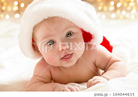 Beautiful little child is celebrating Christmas. New Year's holidays. A child in a Christmas costume. Childhood and people concept - happy Newborn baby in Santa hat over holidays lights background Beautiful little child is celebrating Christmas. New Year's holidays. A child in a Christmas costume. Childhood and people concept - happy Newborn baby in Santa hat over holidays lights background 104044635