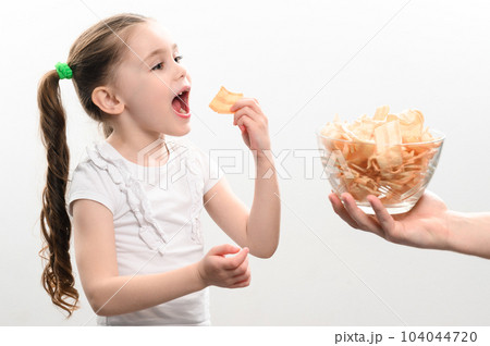 Little girl is given a big bowl of chips snacks with lard, white background portrait of little girl eating chips. 104044720