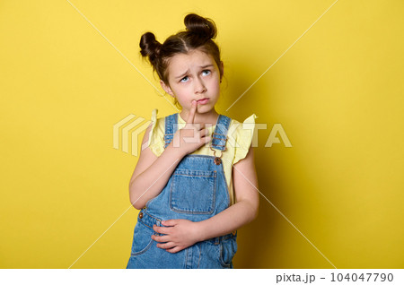 Pensive little kid girl with stylish hairstyle, dressed in yellow t-shirt and blue overalls, thoughtfully looking aside, putting finger on her chin, thinking, reasoning over isolated studio background 104047790