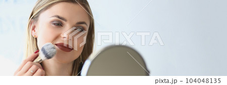 Smiling young woman looks in the mirror and paints herself with blush brushes Smiling young woman looks in the mirror and paints herself with blush brushes 104048135
