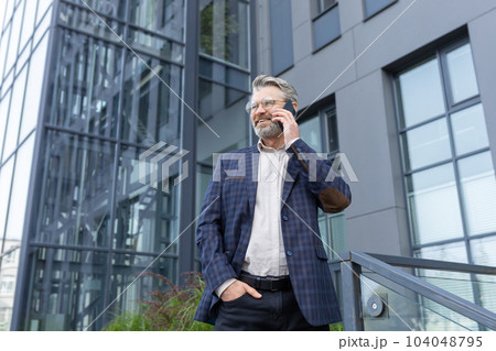 Successful adult mature boss outside office building talking on the phone, gray-haired businessman happy talking with client, man negotiating with business partner. Successful adult mature boss outside office building talking on the phone, gray-haired businessman happy talking with client, man negotiating with business partner. 104048795