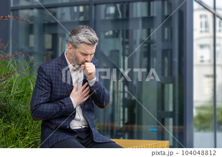 Sick mature man outside office building, adult gray-haired businessman coughing sitting on bench near work, senior boss in business suit with cold virus. 104048812