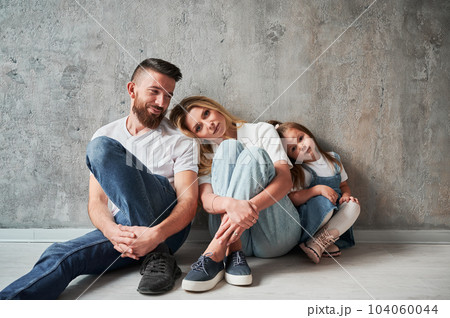 Adorable little girl and parents sitting by the wall in new apartment. Woman leaning head on husband shoulder while resting with family in new home. 104060044