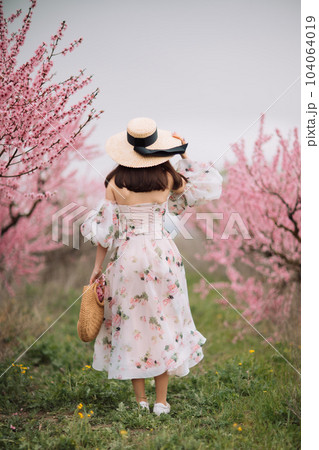 Woman blooming peach orchard. Against the backdrop of a picturesque peach orchard, a woman in a long dress and hat enjoys a peaceful walk in the park, surrounded by the beauty of nature. 104064019