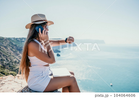 Digital nomad, woman in the hat, a business woman with a laptop sits on the rocks by the sea during sunset, makes a business transaction online from a distance. Freelance, remote work on vacation. Digital nomad, woman in the hat, a business woman with a laptop sits on the rocks by the sea during sunset, makes a business transaction online from a distance. Freelance, remote work on vacation. 104064606