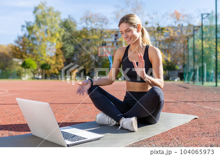 Online training, female trainer sitting on sports mat in stadium at daytime, using laptop for video call for online training, active lifestyle courses. 104065973