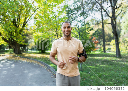Latin American cheerful and smiling running in the park man listening to music and audio podcasts, runner smiling and looking at the camera, joyful sportsman outdoors in the day. 104066352