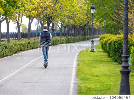 A young guy rides an electric scooter along a beautiful green alley in a city park. 104066464