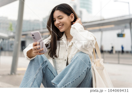 Bright portrait of cheerful young woman 25s with shopper on shoulder waiting public transport at station smiling beautifully and texting Bright portrait of cheerful young woman 25s with shopper on shoulder waiting public transport at station smiling beautifully and texting 104069431