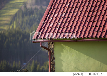 Detail of new modern house cottage corner with stucco walls decorated with natural stones, red shingled roof and rain gutter pipe system on blurred spruce forest copy space background. Detail of new modern house cottage corner with stucco walls decorated with natural stones, red shingled roof and rain gutter pipe system on blurred spruce forest copy space background. 104071540