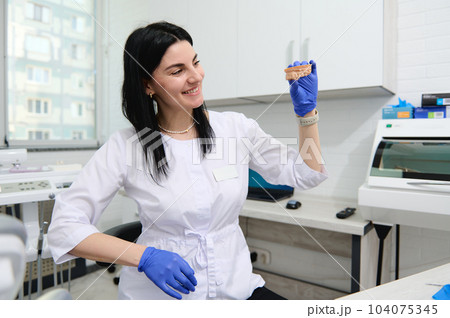 Professional experienced dentist woman, maxillofacial surgeon, prosthetic engineer, dental technician holding plaster cast of a human jaw model with teeth. Installing veneer or prosthetics in clinic 104075345