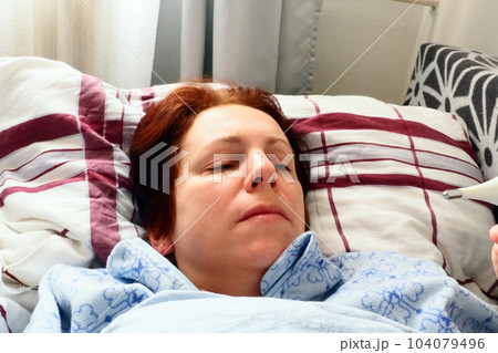Sick woman in bed. Headache, runny nose and cold. Paleness of the face and faintness of the body. Feeling unwell. The patient lies on a pillow, holds a thermometer and measures the temperature Sick woman in bed. Headache, runny nose and cold. Paleness of the face and faintness of the body. Feeling unwell. The patient lies on a pillow, holds a thermometer and measures the temperature 104079496