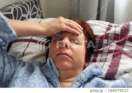 Sick woman in bed. Headache, runny nose and cold. Paleness of the face and faintness of the body. Feeling unwell. The patient holds his forehead with his hand and covers his eyes. 104079527