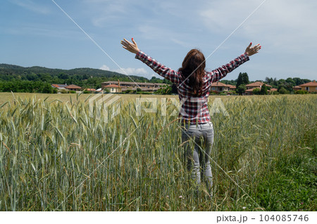 A young woman stands in a wheat field in summer with her back with her arms spread to the sides. 104085746