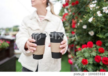 A girl in a residential neighborhood holds cups of coffee. Two glasses of hot drinks in the hands of a young woman. Breakfast on the go. Picnic and going out concept.  104085918